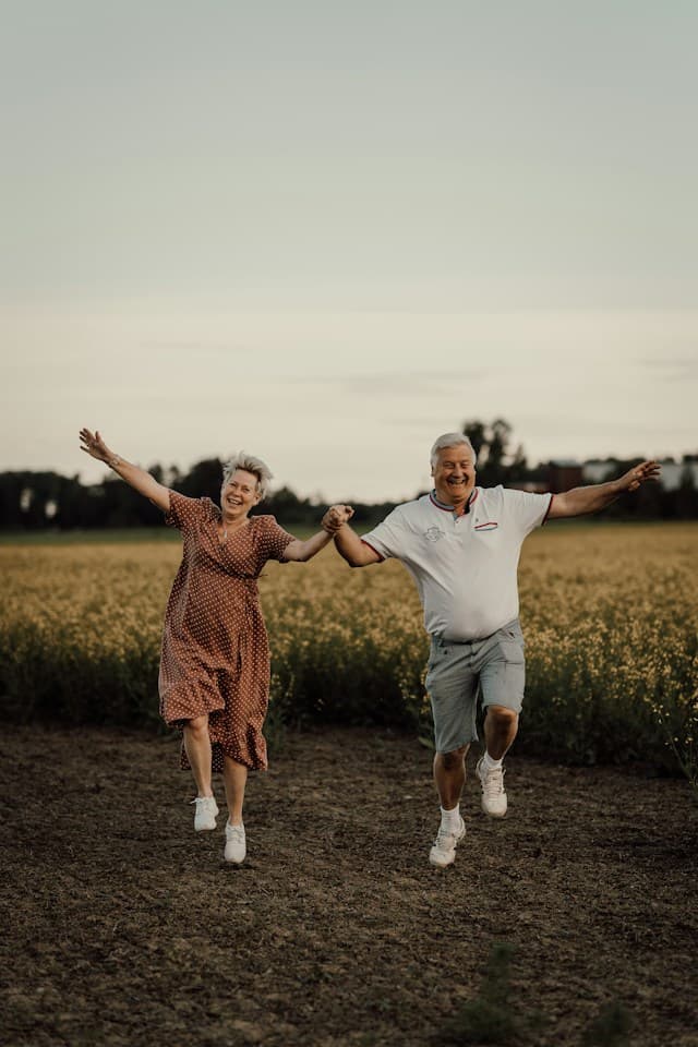 A retired couple laughing and running through a countryside field, holding hands
